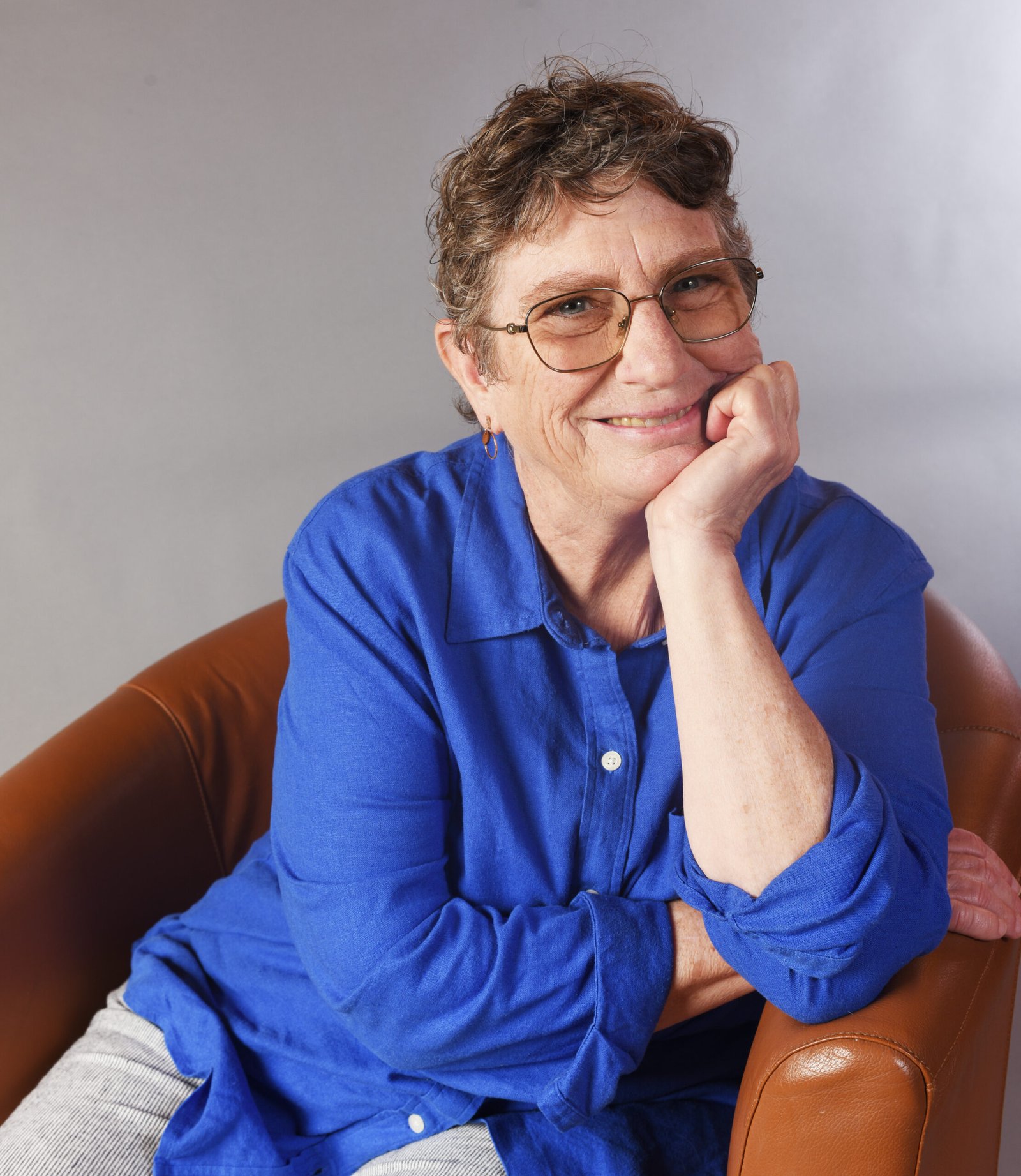 A woman therapist wearing a blue shirt and sitting in a chair with her head in one hand, smiling.