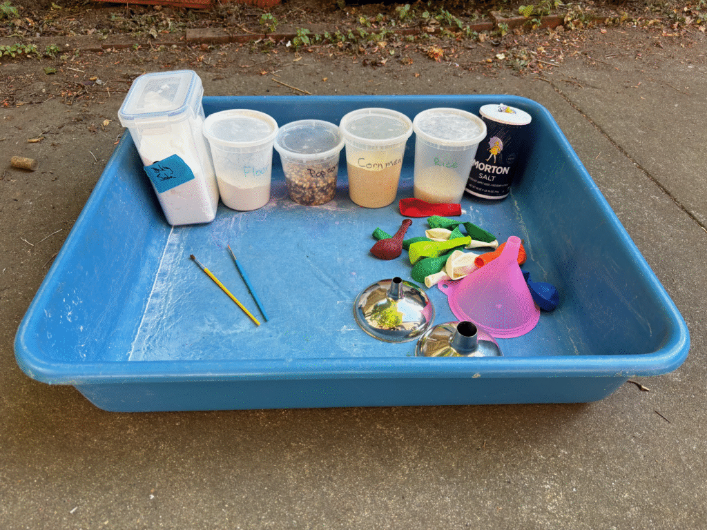 Large flat tub sitting on the ground. At one end are six plastic containers filled with flour, popcorn, cornmeal and rice. A carton of salt is at one end. In front of the containers are a pile of unfilled balloon, three funnels and two thin paint brushes
